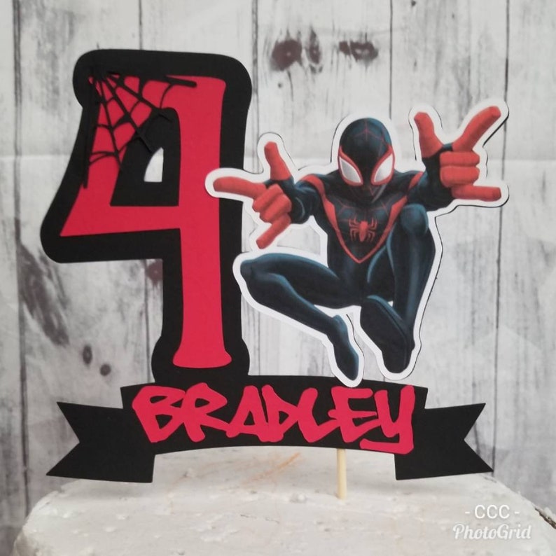 May include: Red and black cake topper featuring a cartoon Spider-Man character with a web design and the number 4. A black banner with the name Bradley in red letters is below the character.