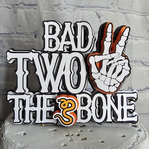 May include: A multi-layered cake topper featuring the text BAD TWO THE BONE in a bold, black-outlined white font. The design includes a white skeleton hand making a peace sign and a small orange snake replacing the letter O in TO.
