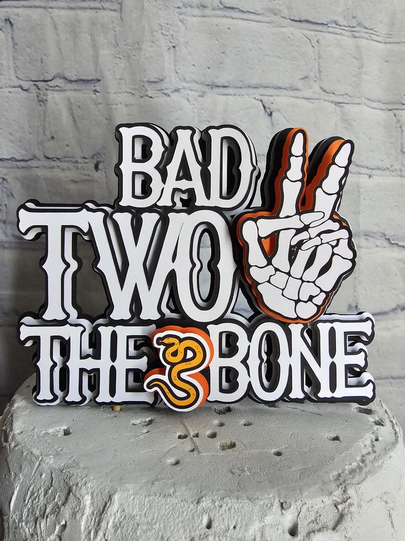 May include: A multi-layered cake topper featuring the text BAD TWO THE BONE in a bold, black-outlined white font. The design includes a white skeleton hand making a peace sign and a small orange snake replacing the letter O in TO.