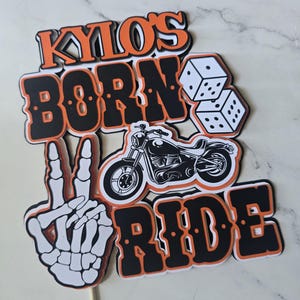 May include: A layered cake topper featuring the text KYLO'S BORN TO RIDE in black and orange lettering. The design includes a white skeleton hand making a peace sign, a black motorcycle illustration, and two white dice with black pips.