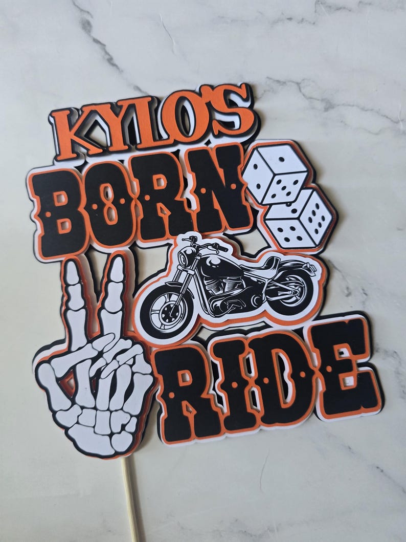 May include: A layered cake topper featuring the text KYLO'S BORN TO RIDE in black and orange lettering. The design includes a white skeleton hand making a peace sign, a black motorcycle illustration, and two white dice with black pips.