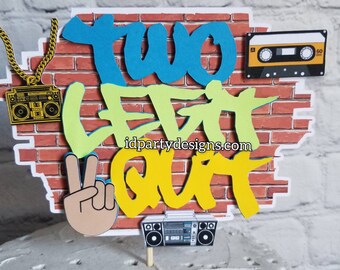 May include: A cake topper with a brick wall background and the words "Two Legit 2 Quit" in graffiti style. The topper also includes a gold chain, a boombox, a cassette tape, and a peace sign.