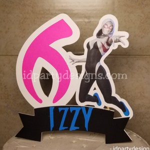May include: A white and pink cake topper featuring a superhero character in a white and black costume with a red spider symbol on the chest. The character is jumping and has a white mask with a red spider symbol. The topper also includes a pink number 6 and a black banner with the name "IZZY" in blue letters.