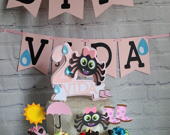 May include: A birthday cake with cupcakes, a banner that says "BIRTHDAY VIDA", and spider and rain-themed decorations. The cake has a number 2 cake topper. The cupcakes have various toppings and decorations.