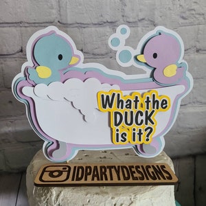 Puede incluir: Un adorno para pastel con dos patos en una ba&ntilde;era, con el texto "What the duck is it?" y el logotipo "IDPartyDesigns".