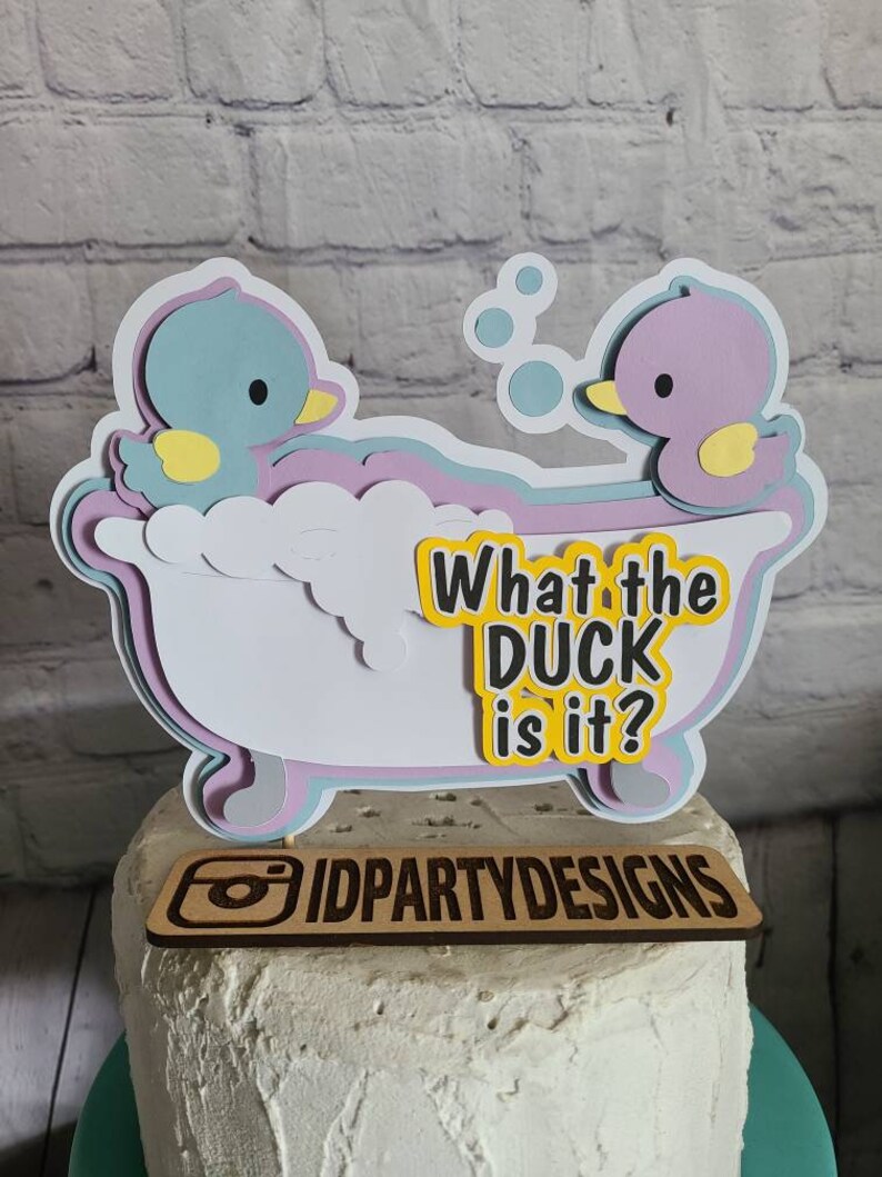Puede incluir: Un adorno para pastel con dos patos en una ba&ntilde;era, con el texto "What the duck is it?" y el logotipo "IDPartyDesigns".