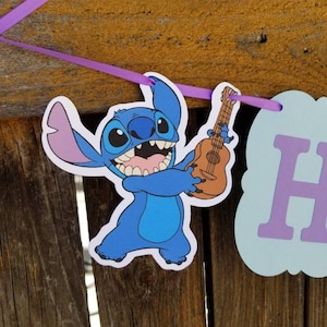 以下が含まれることがあります： ウクレレを持っている漫画のキャラクター、スティッチをフィーチャーしたバナー。キャラクターは青色で、大きな耳と広い笑顔があります。バナーは紫色のリボンでつながれており、水色と紫色の文字が入った看板が含まれています。