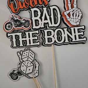 May include: A layered cake topper set featuring the text "Owen's BAD TO THE BONE" in black, white, and orange. The design includes a motorcycle graphic, a skeleton hand making a peace sign, and a separate piece showing dice held by a skeleton hand.