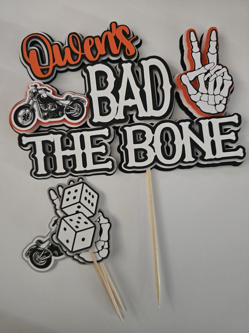 May include: A layered cake topper set featuring the text "Owen's BAD TO THE BONE" in black, white, and orange. The design includes a motorcycle graphic, a skeleton hand making a peace sign, and a separate piece showing dice held by a skeleton hand.