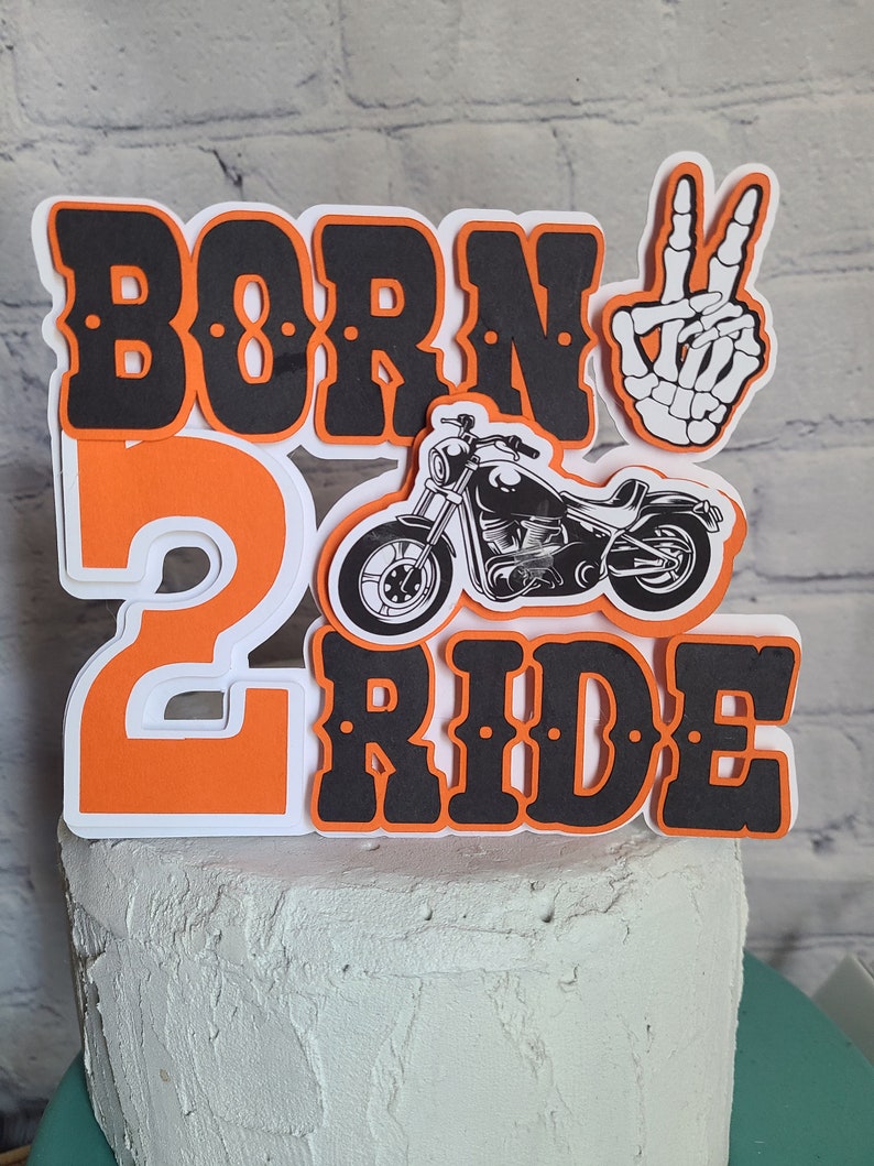 May include: A layered cake topper featuring the text BORN 2 RIDE in black and orange, a black and white motorcycle illustration, and a white skeleton hand making a peace sign. The design is constructed with multiple layers of cardstock.