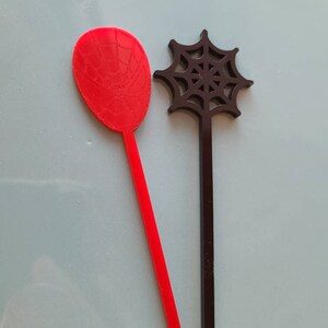 May include: Two plastic stirring sticks with a spider web design. The red stick has a spider web design on the top and the black stick has a spider web design on the top.