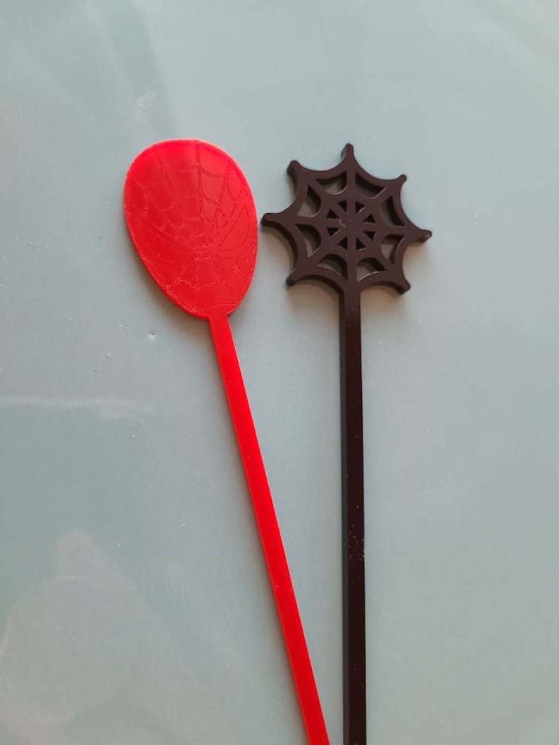 May include: Two plastic stirring sticks with a spider web design. The red stick has a spider web design on the top and the black stick has a spider web design on the top.