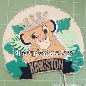 May include: A round cake topper featuring a cartoon lion cub wearing a gold crown. The lion cub is surrounded by green leaves. The text "idpartydesigns.com" and "KINGSTON" are visible on the topper.