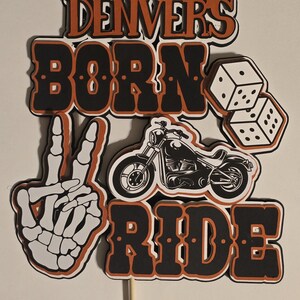May include: A layered cake topper featuring the text "DENVER'S BORN TO RIDE" in black and orange lettering. The design includes a white skeleton hand making a peace sign, a black motorcycle illustration, and two white dice with black dots.