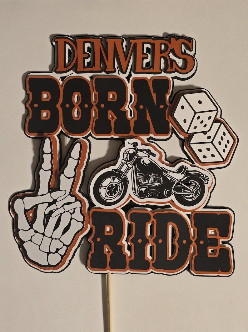 May include: A layered cake topper featuring the text "DENVER'S BORN TO RIDE" in black and orange lettering. The design includes a white skeleton hand making a peace sign, a black motorcycle illustration, and two white dice with black dots.