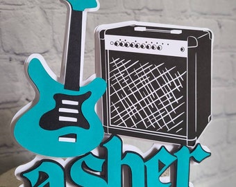 Puede incluir: Un adorno para pastel de guitarra y amplificador azul turquesa con el nombre "asher" en letras azul turquesa.
