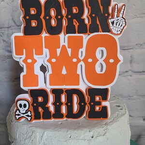 May include: A layered paper cake topper featuring the text BORN TWO RIDE in bold orange and black lettering. The design includes a white skeleton hand making a peace sign and a small white skull and crossbones icon on a white background.