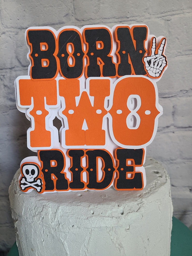 May include: A layered paper cake topper featuring the text BORN TWO RIDE in bold orange and black lettering. The design includes a white skeleton hand making a peace sign and a small white skull and crossbones icon on a white background.