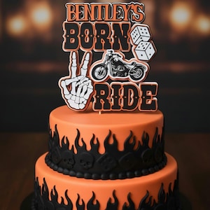 May include: A two-tier orange cake decorated with black flame and skull motifs, topped with a custom cake topper featuring the text BENTLEY'S BORN TO RIDE, a black motorcycle illustration, a pair of white dice, and a skeleton hand peace sign.