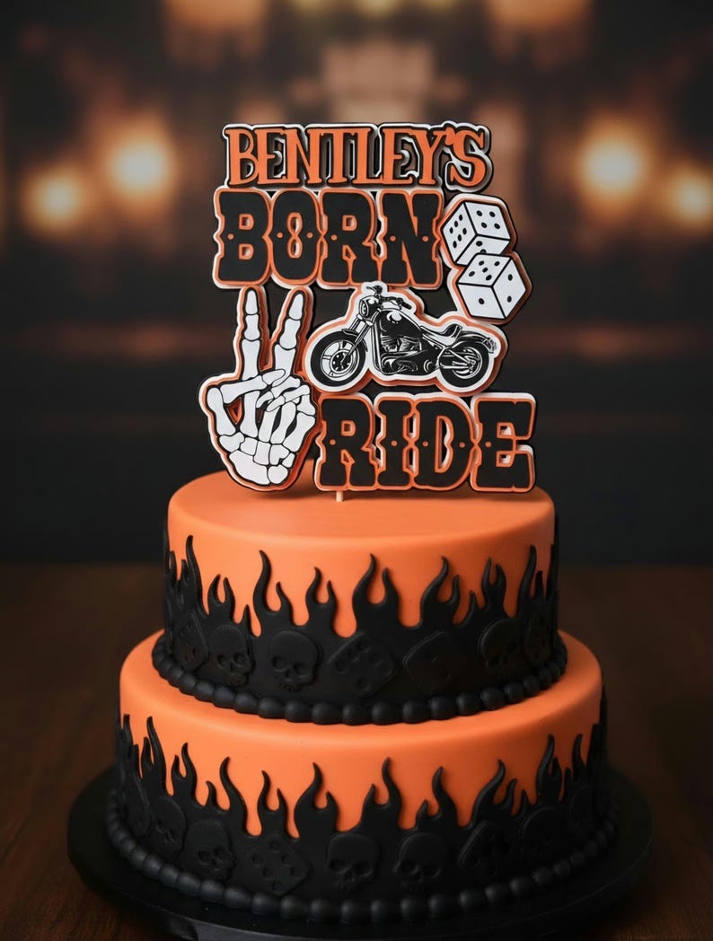 May include: A two-tier orange cake decorated with black flame and skull motifs, topped with a custom cake topper featuring the text BENTLEY'S BORN TO RIDE, a black motorcycle illustration, a pair of white dice, and a skeleton hand peace sign.