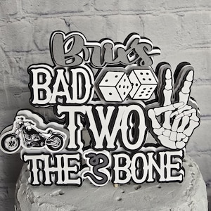May include: A multi-layered cake topper featuring the text "BRU'S BAD TWO THE BONE" in black, white, and gray. The design includes a black motorcycle, two white dice, and a white skeletal hand making a peace sign, all mounted on a gray cake.