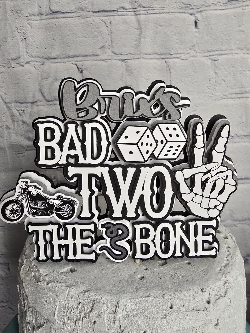 May include: A multi-layered cake topper featuring the text "BRU'S BAD TWO THE BONE" in black, white, and gray. The design includes a black motorcycle, two white dice, and a white skeletal hand making a peace sign, all mounted on a gray cake.