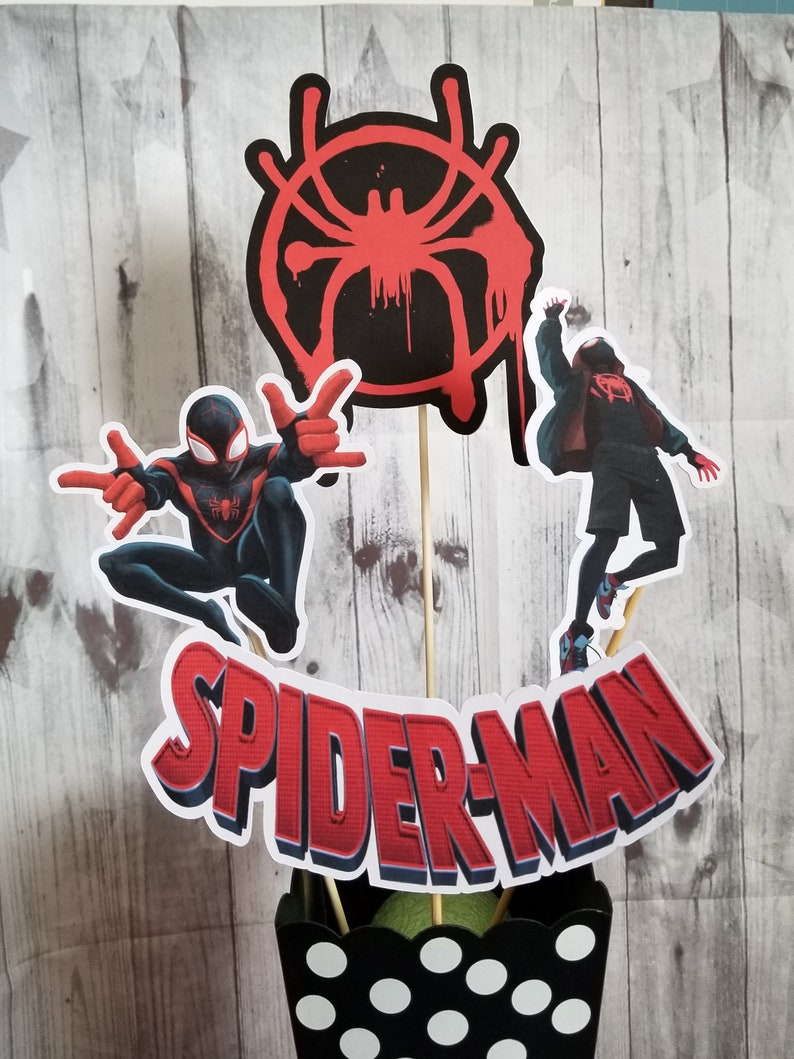 May include: A black and red Spider-Man centrepiece with a black and white polka dot vase. The centrepiece features a red Spider-Man symbol, a red and black Spider-Man character, and a red and black Spider-Man character in a red jacket. The text "SPIDER-MAN" is in red with a blue outline.