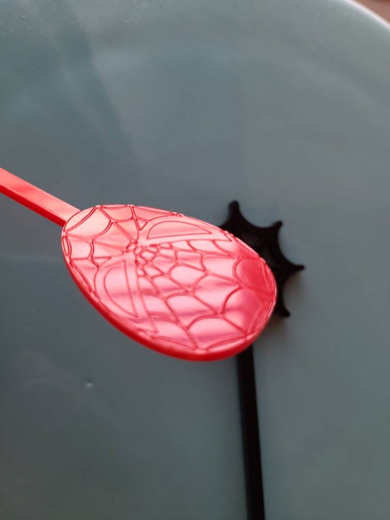 May include: Red plastic spoon with a spider web design. The spoon is part of a set of Spider-Man themed party supplies.