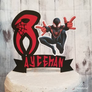 May include: Black and red cake topper featuring a cartoon of Miles Morales, also known as Spider-Man, in a superhero pose. The topper also includes a red number 8 with a black spider web design and a black banner with the name "AYCEHAN" in red letters.