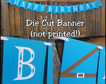 May include: A blue die-cut banner with white text spelling "HAPPY BIRTHDAY" strung across a wooden fence. The banner has brown accents and white ribbon. The text "Die Cut Banner (not printed!)" is also visible.
