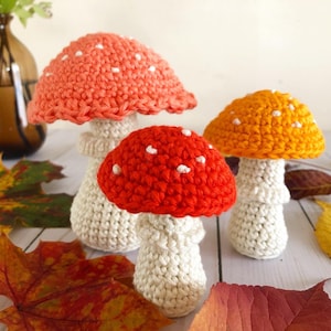May include: Three handmade crochet mushrooms in shades of red, orange, and peach, each with white polka dots on the caps. The stems are a cream color. Autumn leaves are scattered around the mushrooms, creating a fall theme.