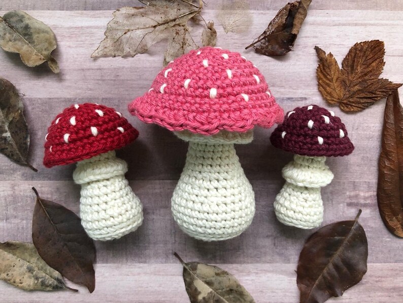 May include: Three handmade crochet mushrooms in varying sizes. The mushroom caps are red, pink, and burgundy, each with white spots. The stems are off-white. The mushrooms are arranged on a wooden surface with dried leaves.