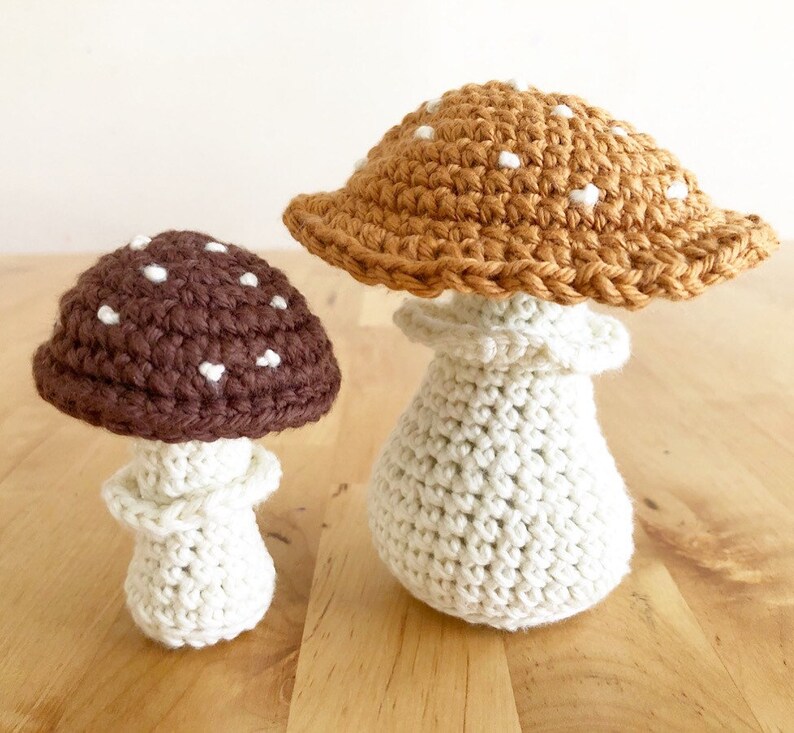 May include: Two crocheted mushroom decorations. One mushroom has a brown cap with white spots and a cream-colored stem. The other mushroom has a tan cap with white spots and a cream-colored stem. Both are handmade.