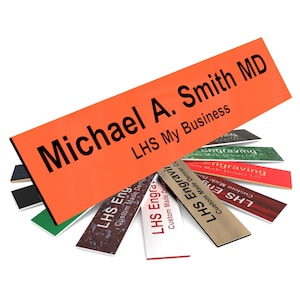 May include: A close-up of a bright orange rectangular nameplate with black text that reads "Michael A. Smith MD" and "LHS My Business". The nameplate is surrounded by several smaller nameplates with different colors and designs.