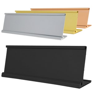 May include: Four desk nameplates in different colors: silver, gold, copper, and black. The nameplates are rectangular with a slight angle at the base for stability.