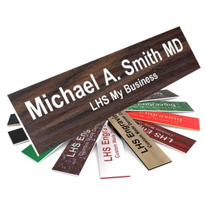 May include: A wooden nameplate with the text "Michael A. Smith MD" and "LHS My Business" in white lettering. The nameplate is surrounded by several smaller nameplates with the text "LHS Engraving" and "Custom Made Denver Co."