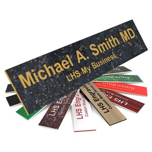 May include: A black and gold engraved nameplate with the text "Michael A. Smith MD" and "LHS My Business". The nameplate is surrounded by smaller, colorful engraved nameplates with the text "LHS Engraving" and "Custom Made Denver CO".