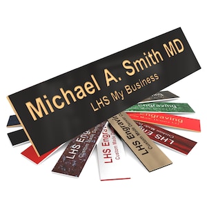 May include: A black nameplate with gold lettering that reads "Michael A. Smith MD" and "LHS My Business." Several other nameplates with various colors and text are fanned out below.
