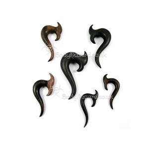 May include: Set of six wooden ear gauges in black and brown colors. The gauges are shaped like a curved hook with a pointed end.