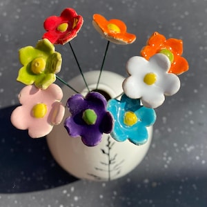 Könnte beinhalten: Ein Strauß bunter Keramikblumen in einer weißen Vase mit schwarzem Design. Die Blumen sind in den Farben Rot, Orange, Gelb, Grün, Rosa, Lila und Blau.