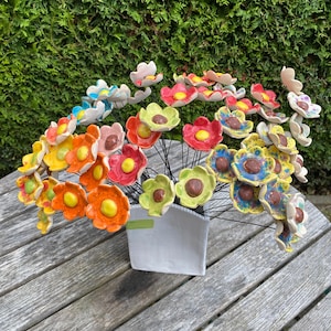 Könnte beinhalten: Ein Strauß bunter Keramikblumen in einer weißen Vase. Die Blumen sind in Rot-, Orange-, Gelb-, Grün-, Blau- und Weißtönen gehalten. Die Vase ist ein schlichtes, modernes Design.