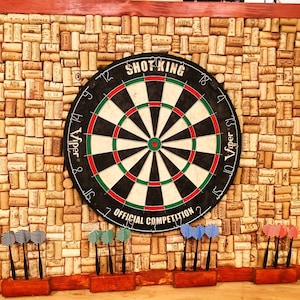 May include: A black and white dartboard with a red bullseye, surrounded by cork tiles. The dartboard has the text "Shot King" and "Viper" printed on it. The text "Official Competition" is printed below the bullseye. Six darts are stuck in the dartboard.