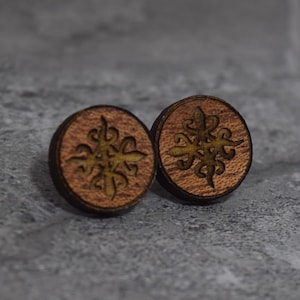May include: Two round wooden earrings with a decorative floral design. The earrings are brown with a lighter brown floral pattern in the center. The earrings are on a gray surface.