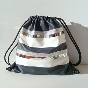 Puede incluir: Una bolsa de cordón de rayas blancas y negras con detalles de purpurina plateada. La bolsa está hecha de un material de imitación de cuero.