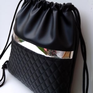 Può includere: Zaino nero con coulisse, con una sezione inferiore trapuntata e una sezione superiore liscia. La borsa presenta un accento argentato e cordini neri. Il design è semplice e moderno, adatto a vari usi.