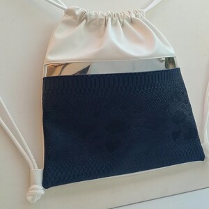 Op de afbeelding: Een witte trekkoordtas met een blauw slangenhuidpatroon op de onderste helft. De tas heeft een zilveren band die de twee kleuren scheidt.