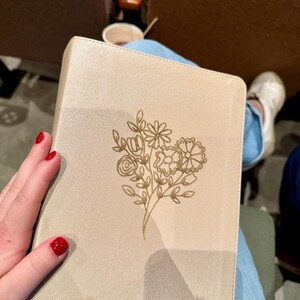 May include: A beige book with a gold floral design. The cover features an embossed bouquet of flowers. The book is held by a person wearing blue jeans and white sneakers. The person has red nail polish.
