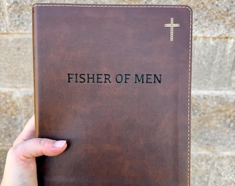 May include: A brown leather-bound book with the text "FISHER OF MEN" in black lettering. A gold cross is in the upper right corner. The book is held in a person's hand.