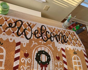 May include: A rectangular doormat featuring a gingerbread house design with the word Welcome in black script. The mat includes white icing details, red and white candy cane pillars, heart accents, and a green wreath on the door.