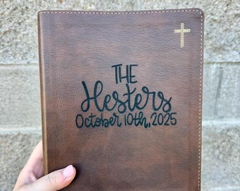 May include: A brown leather-bound book with the words "THE Hesters October 10th, 2025" in black script. A gold cross is in the upper right corner. The book has a stitched border.